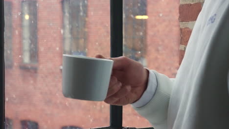 person sips a hot drink by the window as snow falls, cozy and serene