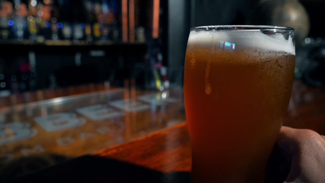 Una mano appoggia un bicchiere di birra artigianale su un bancone da bar in legno.