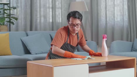 지친 아시아 남성 집주인이 집에서 스프레이로 테이블을 청소하고 있습니다.