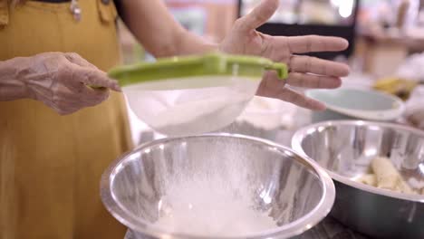 mujer madura tamizando harina en un cuenco de metal en la cocina