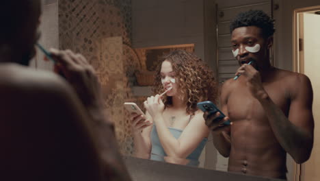 young couple using phones and brushing teeth in front of bathroom mirror