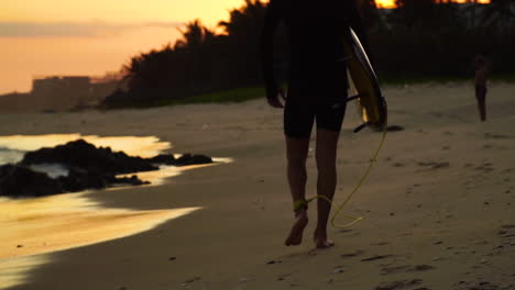 slow mo, surfer guy in wetsuit holding surfboard walking on beach during golden hour