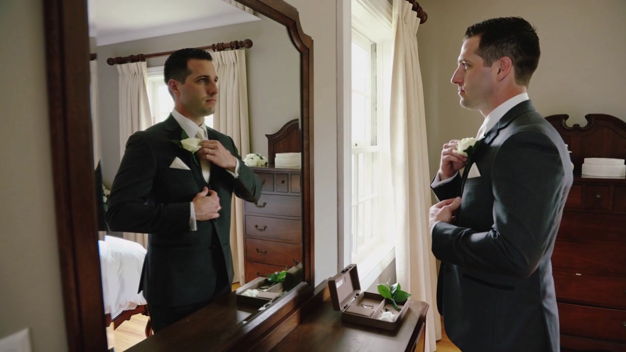 Buttoning dark suit jacket, man adjusting tie, checking white boutonniere at mirror before wedding