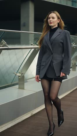 businesswoman in a gray blazer walks confidently along a modern office terrace, holding a tablet and appearing focused