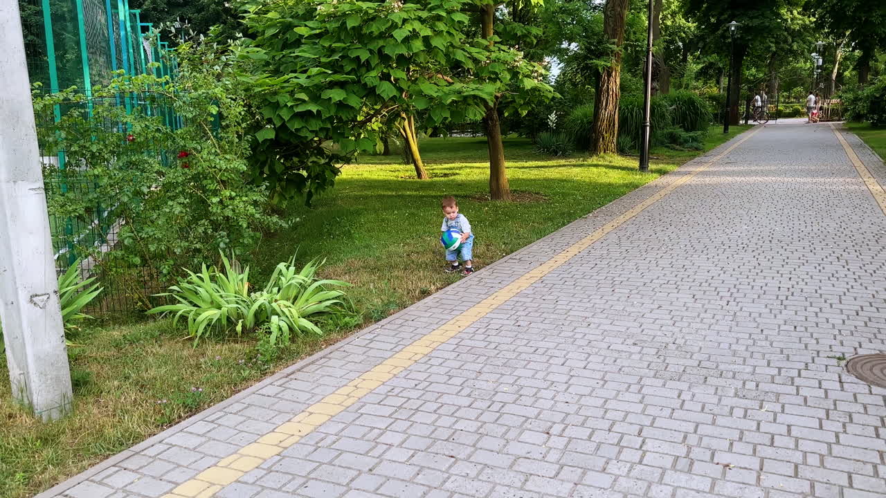 Baby boy goes to pick up the ball from the grass. Kid brings his ball to the camera. Toddler playing in the park.