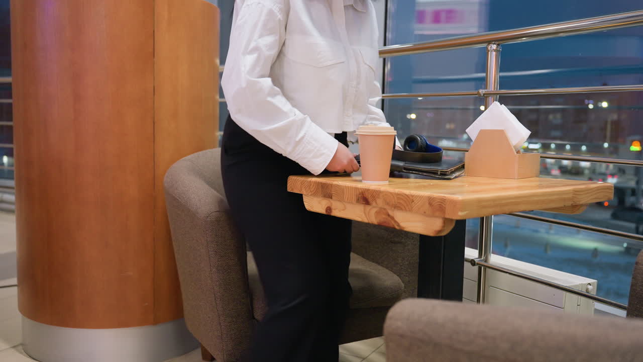 Close up of cozy restaurant seating with woman approaching holding coffee cup, preparing to sit while gently placing coffee and gadget on table near window with night city lights in background