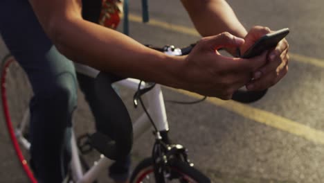 Afroamerikanischer-Mann-In-Der-Stadt,-Der-Mit-Dem-Smartphone-Auf-Dem-Fahrrad-Auf-Der-Straße-Sitzt