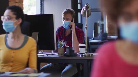 Kaukasischer-Mann-Mit-Gesichtsmaske,-Der-Im-Modernen-Büro-Auf-Dem-Smartphone-Spricht