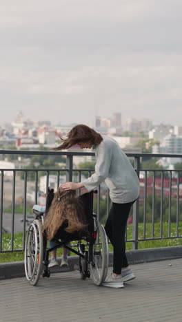 mother puts hand on head of daughter sitting in wheelchair. woman and little girl with spinal cord injury enjoy city view from observation deck