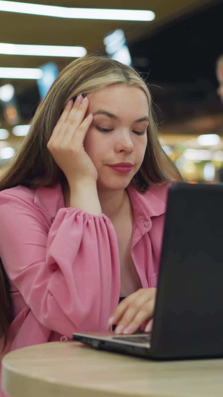dama de vestido rosa sentada sola en un restaurante ocupado quita su mano de su barbilla y trabaja en su portátil, parece aburrida mientras la gente en el fondo se mueve, algunos están sentados