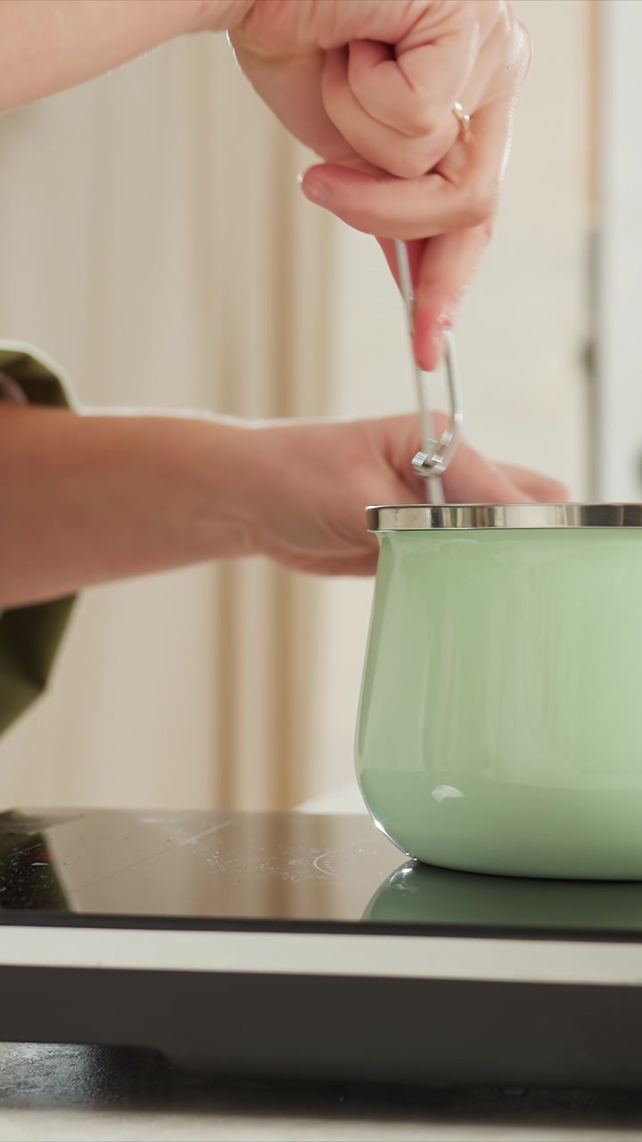 Close up of person stirring and lifting green ceramic pot. Green sleeve visible on arm hands performing continuous. Neutral background suggests indoor setting with vertical lines