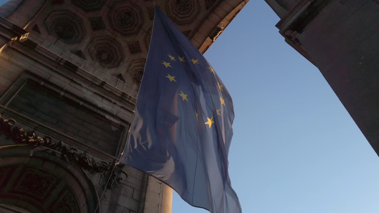 The EU flag waves gently beneath the Cinquantenaire monument in Brussels, highlighted by golden light and set against a clean blue sky. The image captures European unity and historical grandeur
