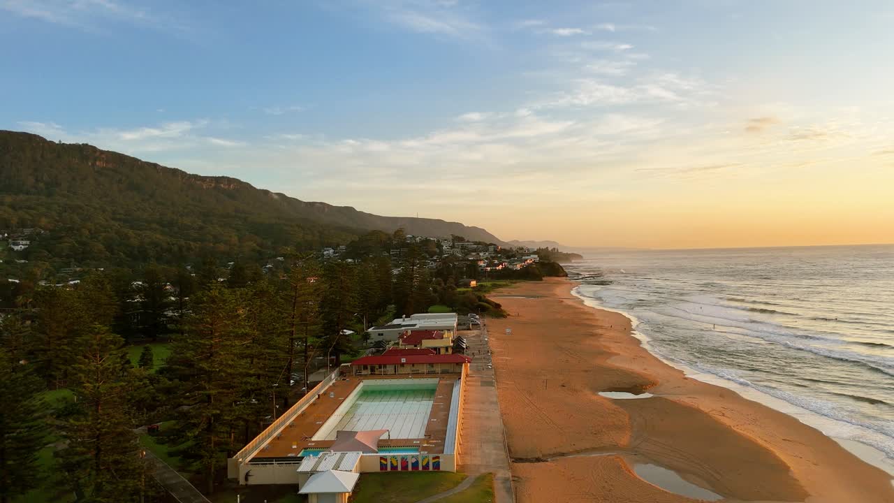 Aerial ascend overview of Thirroul beach and coastal strip with waves rolling onto golden sand, pool along pine trees at sunrise