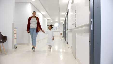 african american mother and daughter in hospital gown holding hands walking in corridor, slow motion