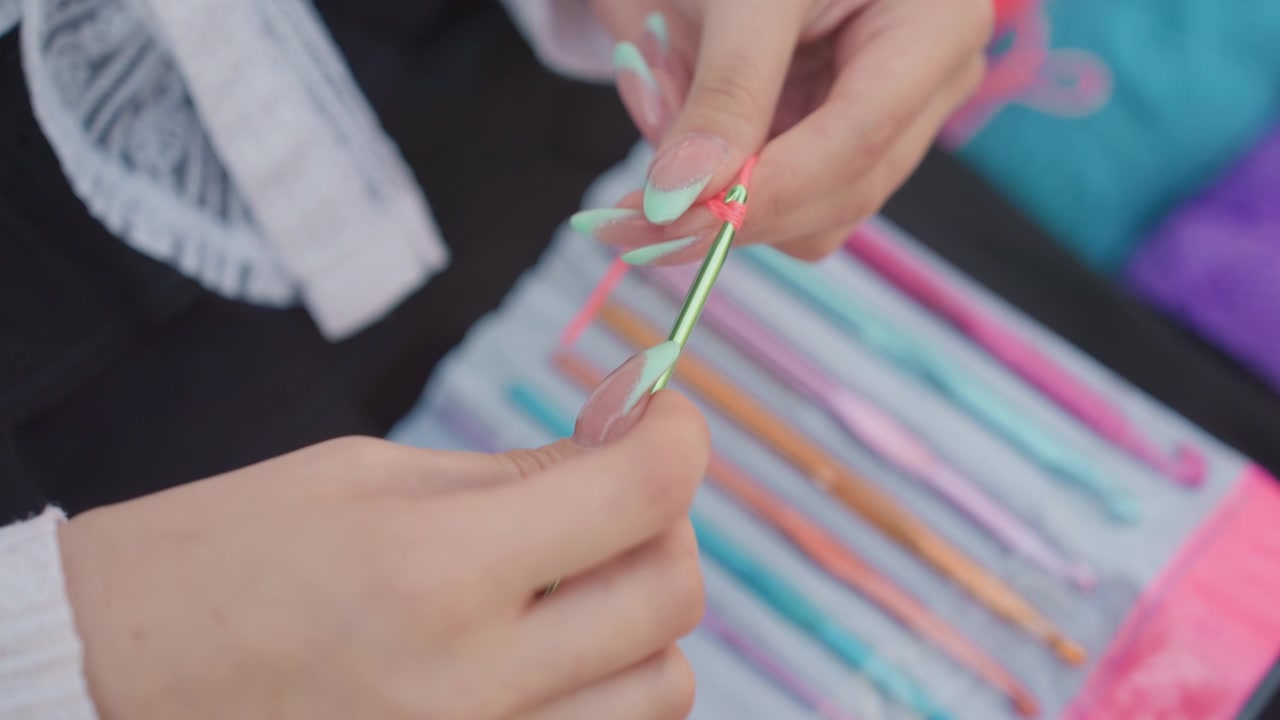 Primo piano di mani con unghie verde menta che lavorano all'uncinetto un anello usando filato rosa e un uncinetto di metallo. Vari uncinetti colorati sono visibili sullo sfondo su un tavolo, con un'illuminazione soffusa che evidenzia i dettagli della mano.