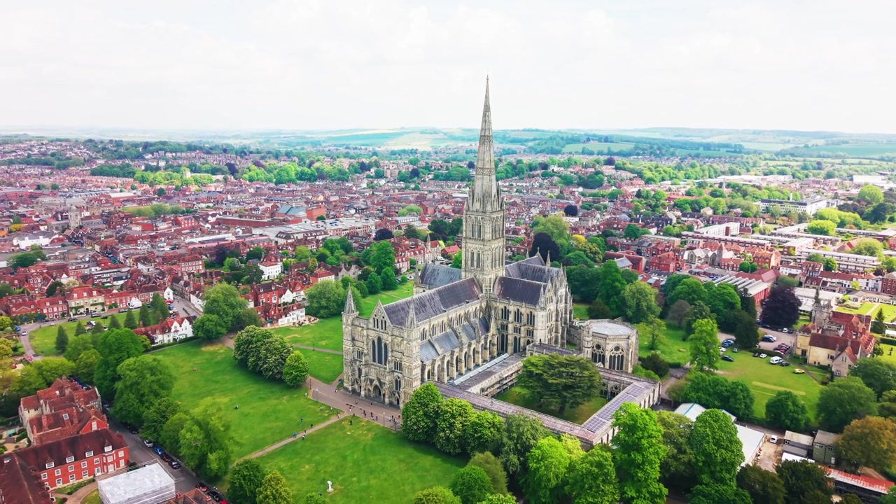 Imagens aéreas de drone da Catedral de Salisbury em Wiltshire, mostrando a icônica torre da catedral, Cathedral Close, o centro histórico da cidade, telhados, área verde de parque e a paisagem rural circundante.