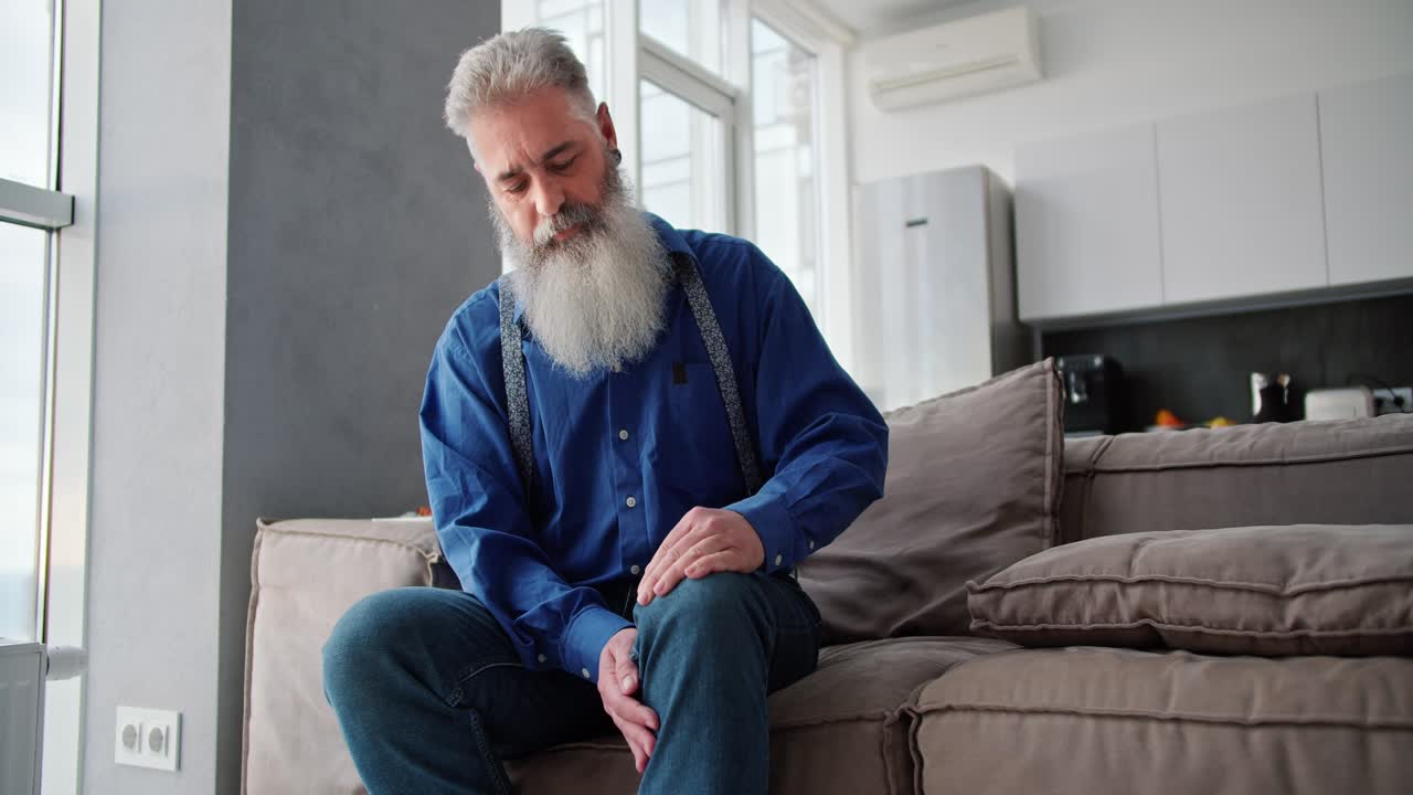 An elderly man with gray hair and a lush beard in a blue shirt strokes and massages his knee while sitting on a brown modern sofa in an apartment