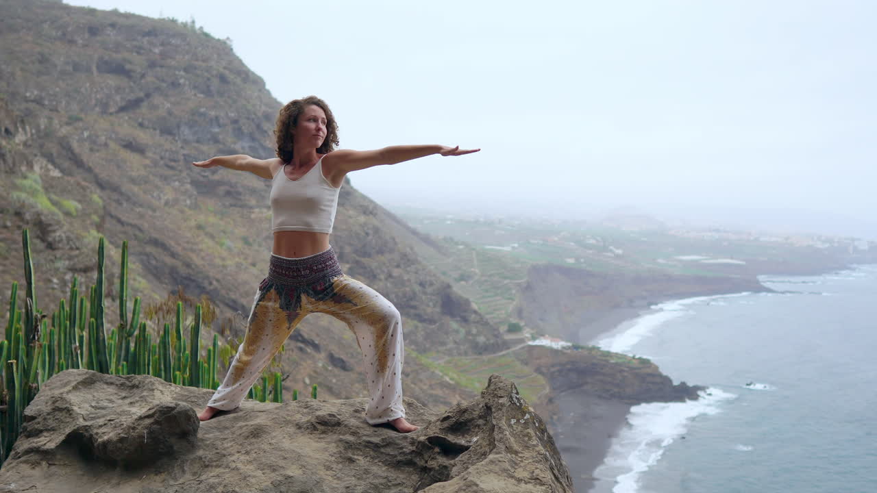 Against the scenic backdrop of the ocean, beach, and rock mountains, a woman's yoga warrior pose signifies motivation, inspiration, and a commitment to outdoor fitness