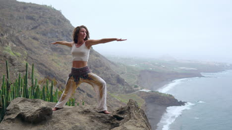 Against-the-scenic-backdrop-of-the-ocean,-beach,-and-rock-mountains,-a-woman's-yoga-warrior-pose-signifies-motivation,-inspiration,-and-a-commitment-to-outdoor-fitness
