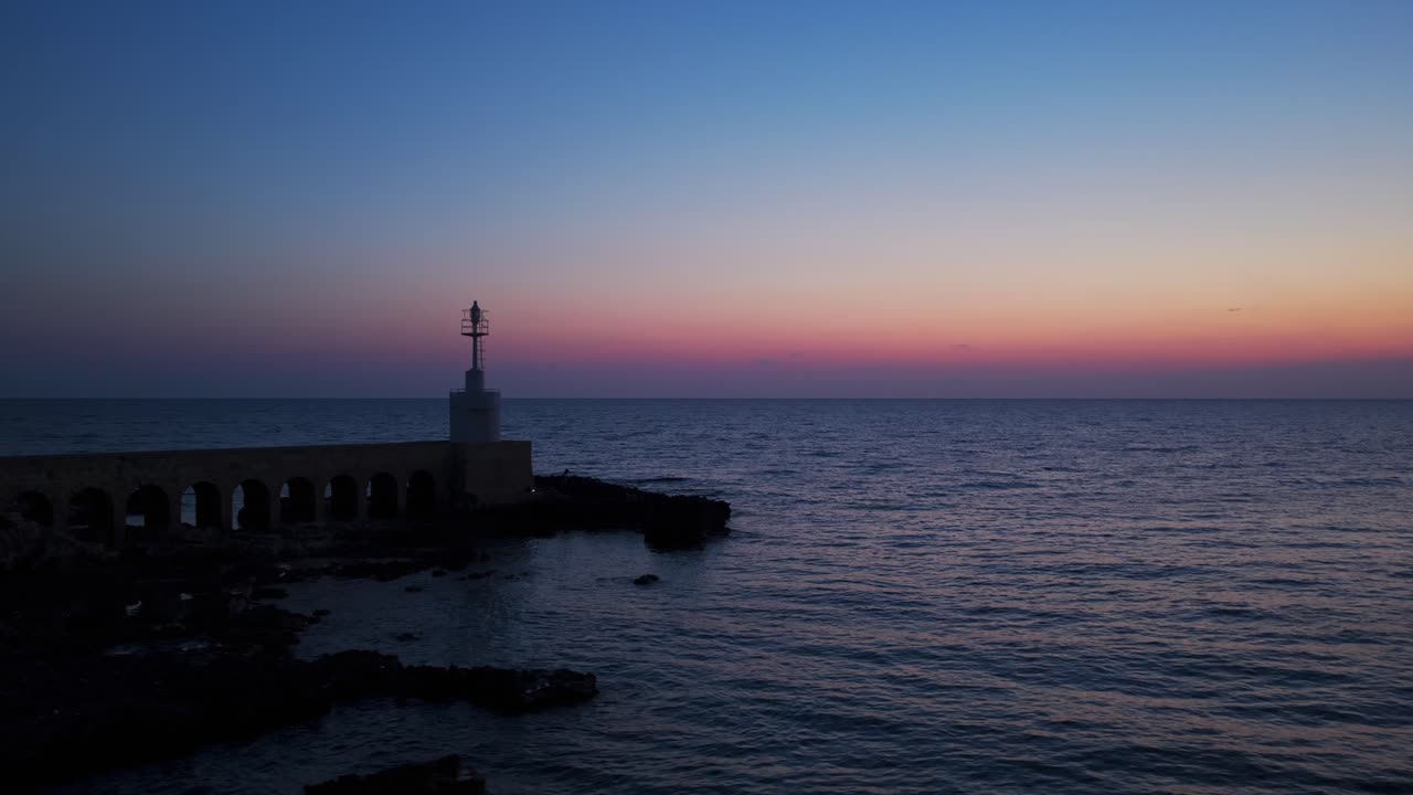 Aerial drone view of Otranto lighthouse silhouetted against colorful twilight sky, calm sea reflects beautiful colors, Puglia, Italy