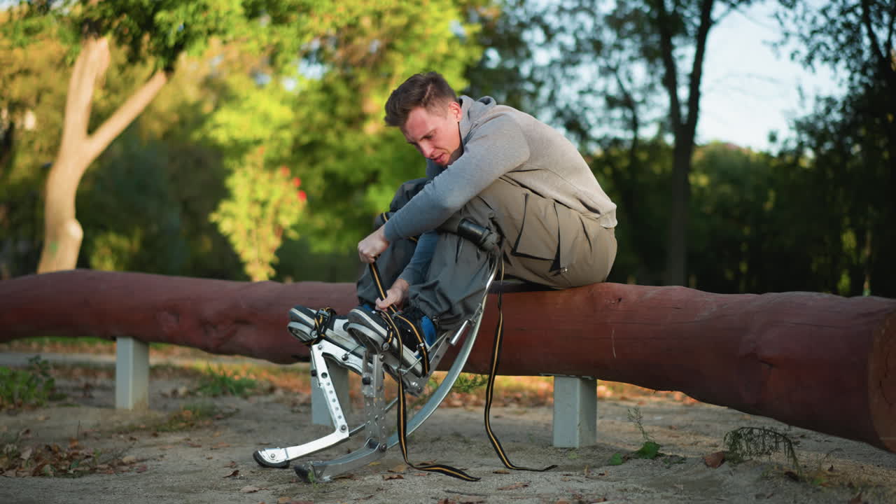Guy sitting on log in park, wearing gray long-sleeved shirt and cargo pants, focused on adjusting spring stilt straps attached to feet, surrounded by green trees, with low camera angle