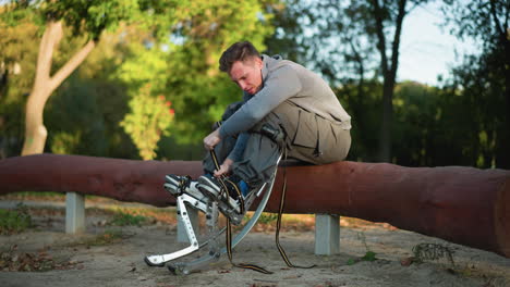 guy sitting on log in park, wearing gray long-sleeved shirt and cargo pants, focused on adjusting spring stilt straps attached to feet, surrounded by green trees, with low camera angle