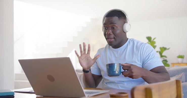 Hombre africano de mediana edad respondiendo a una alerta en su computadora portátil en casa con auriculares, escribiendo y hablando