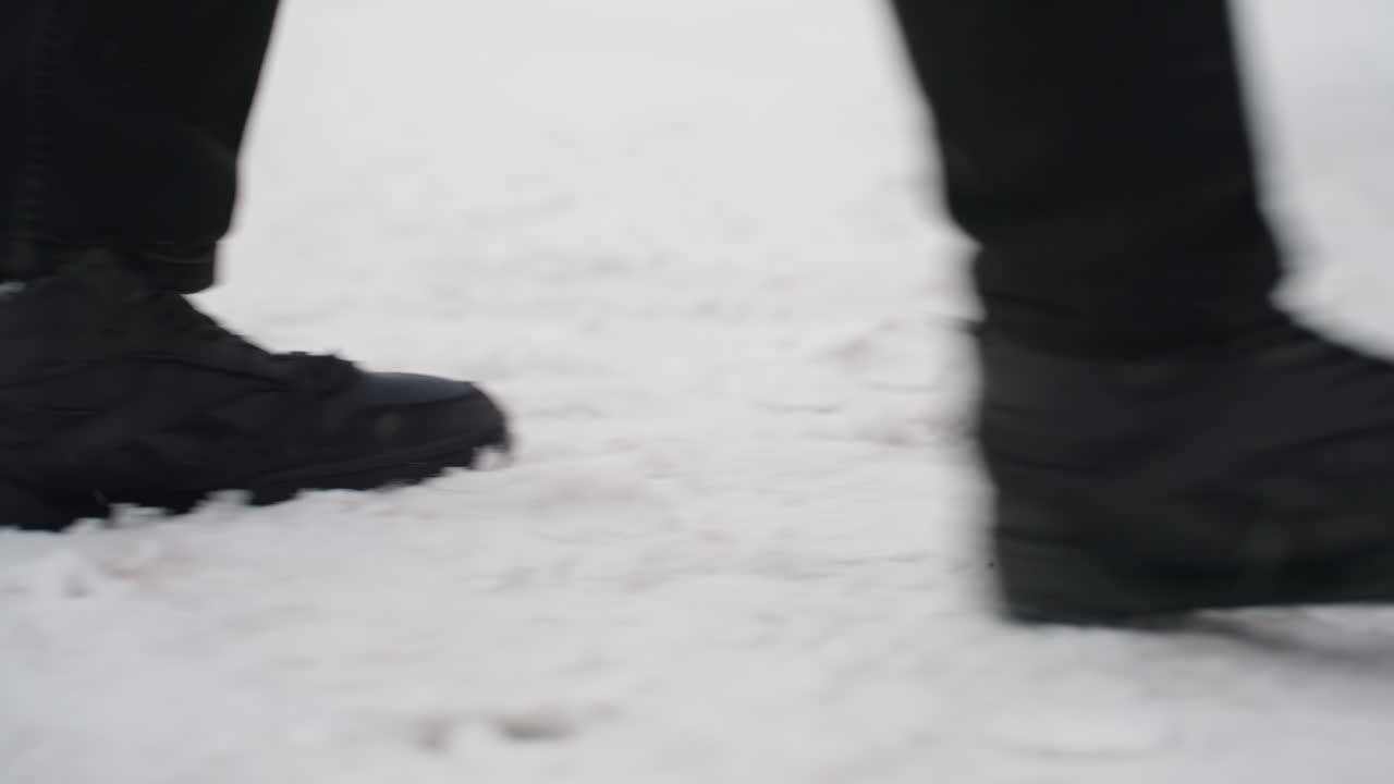 Close up side view of person wearing black boots walking across snow covered ground toward car door opening with flakes falling and parked cars visible in background during cold winter day