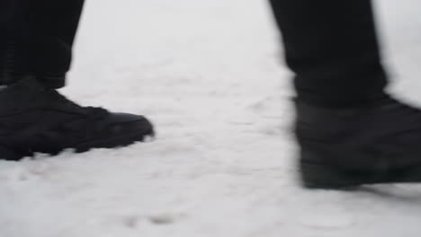 close up side view of person wearing black boots walking across snow covered ground toward car door opening with flakes falling and parked cars visible in background during cold winter day