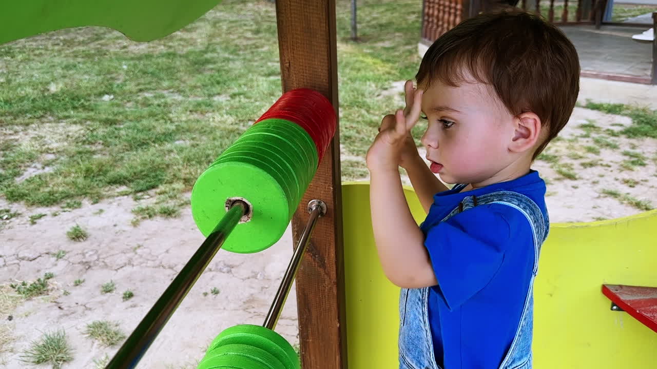 Beautiful Caucasian dark-haired baby in blue t-shirt and jeans romper stands near the abacus. Little boy on the playground.