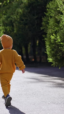 following the happy kid running by the road. baby boy in yellow suit and cap outdoors on sunny weather. vertical video