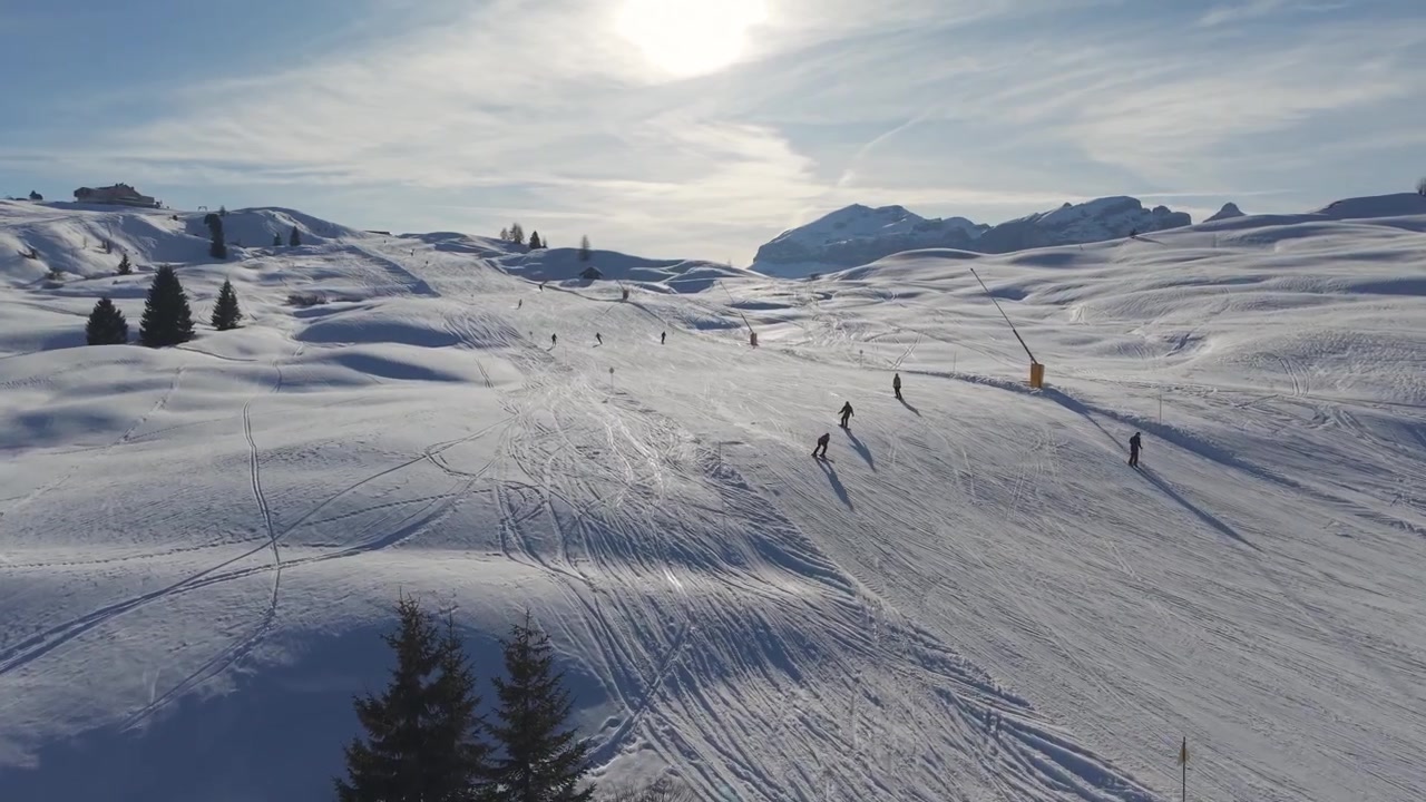 Sunlit ski slope across the Piz Sorega–Pralongià plateau in Alta Badia, with skiers crossing rolling snowfields beneath distant Dolomite peaks.