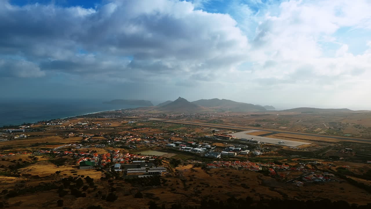 A plain with a cozy town with airport situated on. Silhouettes of mountains are in haze at backdrop. Dramatic cloudscape covering the sky above the Madeira Islands, Portugal.