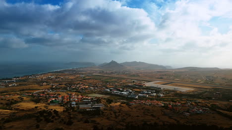 a plain with a cozy town with airport situated on. silhouettes of mountains are in haze at backdrop. dramatic cloudscape covering the sky above the madeira islands, portugal.