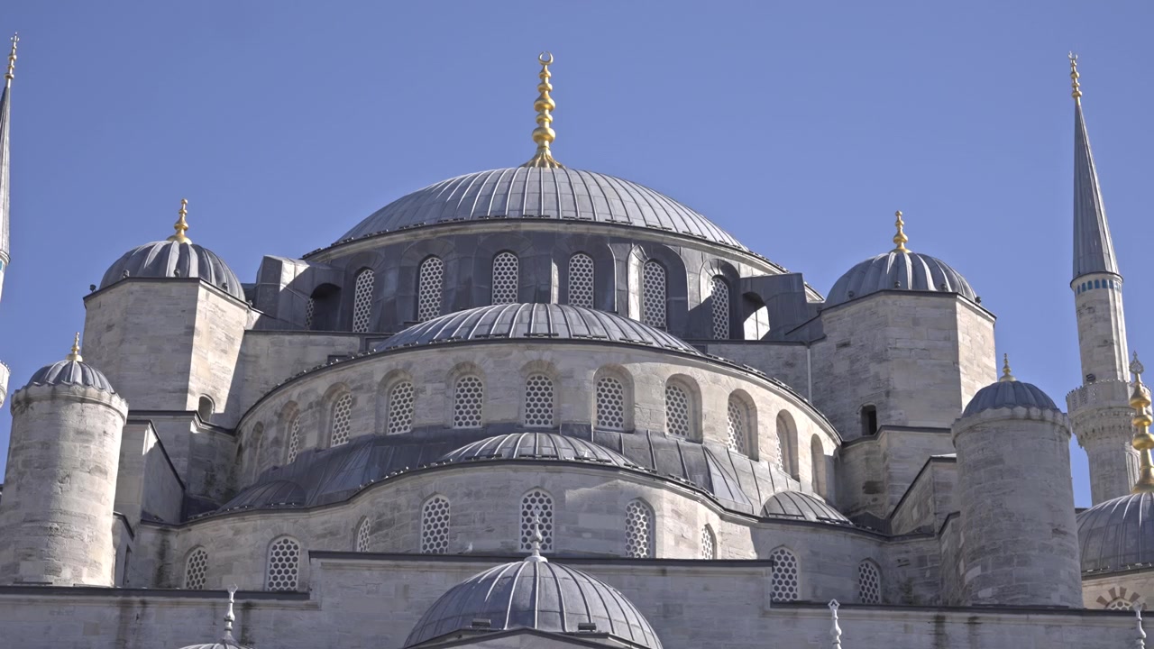 Sultan Ahmet Camii (Mavi Cami) dış cephesinin, basamaklı kubbelerini, altı minaresini ve kemerli avlusunu sergileyen sinematik 4K görüntüler. Açık mavi gökyüzü, İstanbul, Türkiye.