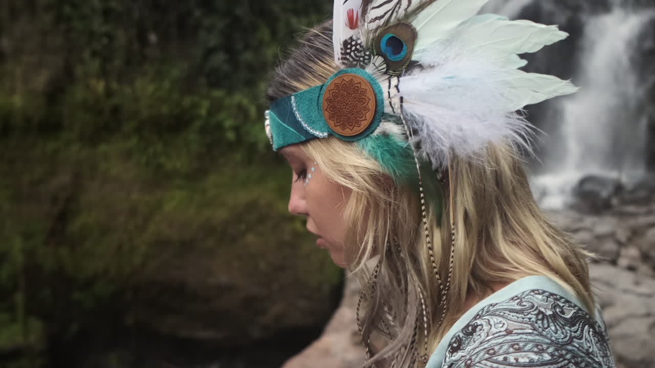una mujer con una gorra de plumas junto a una cascada.
