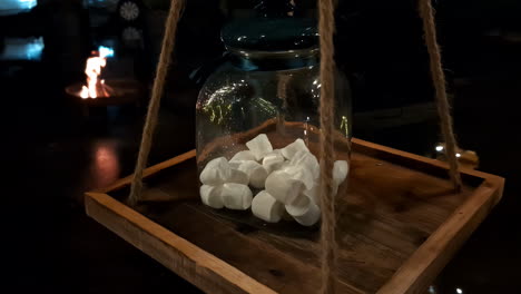 Close-up-shot-of-marshmallows-in-a-glass-jar-with-fire-burning-in-the-background-in-a-dark-room