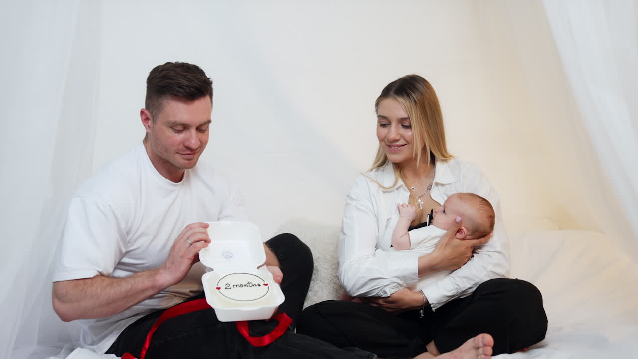 Family of three celebrating the baby second month of life. Happy parents sit together and dad opens the box with a cake.