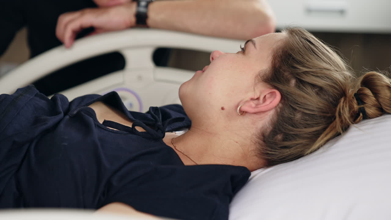 Fair-haired Caucasian woman lies in the hospital bed. Pregnant lady closes eyes as she feels pain from contractions. Close up.