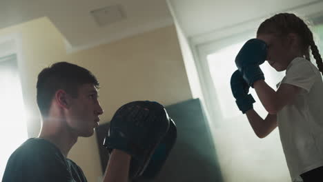 instructor trains little child to punch in gym. friendly trainer adjusts girl fists to right position for boxing in sports club slow motion. physical activity