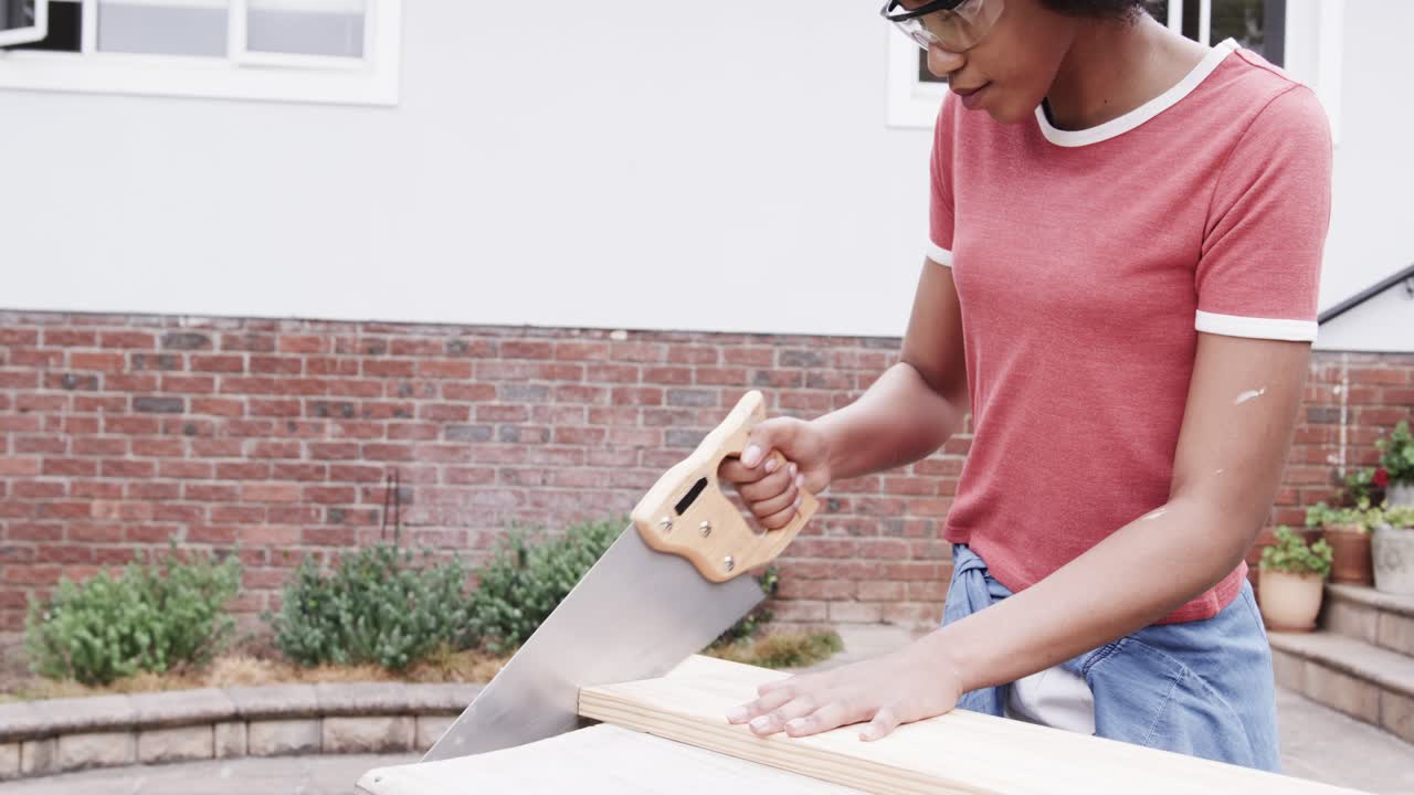 Donna afroamericana che tiene fermo il legno, indossando occhiali di protezione, segando con una sega per il fai da te, nel patio, spazio per il testo