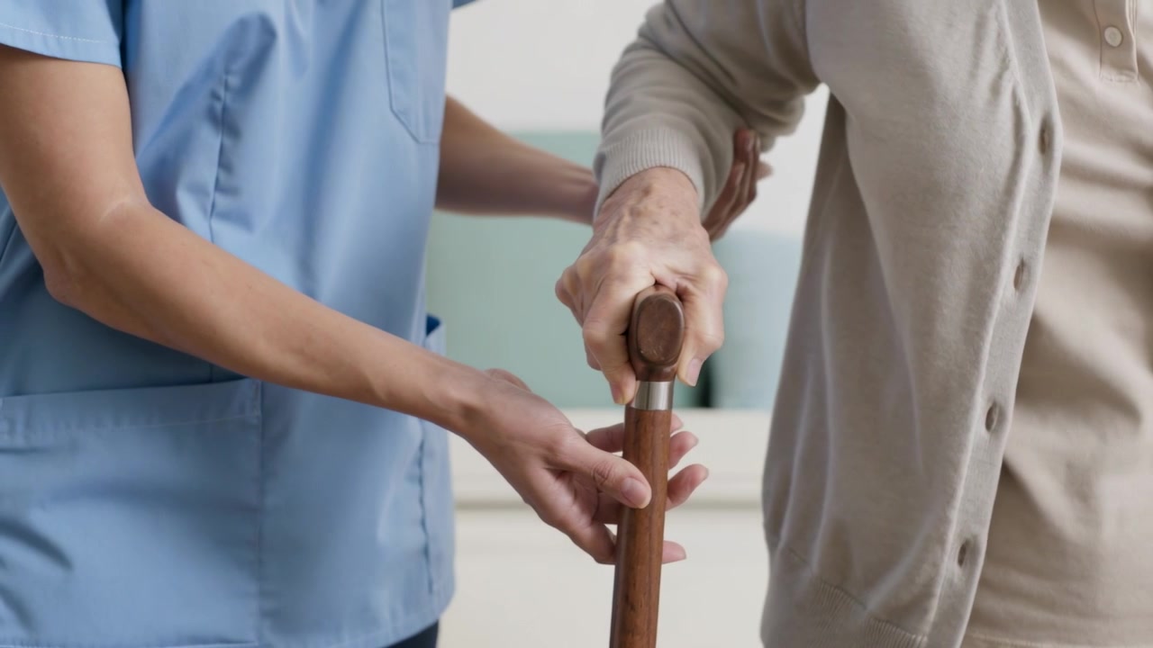 Assistente sanitario in divisa che si protende per guidare un anziano in cardigan con bastone di legno a casa, aiutandolo a mantenere l'equilibrio per alzarsi.