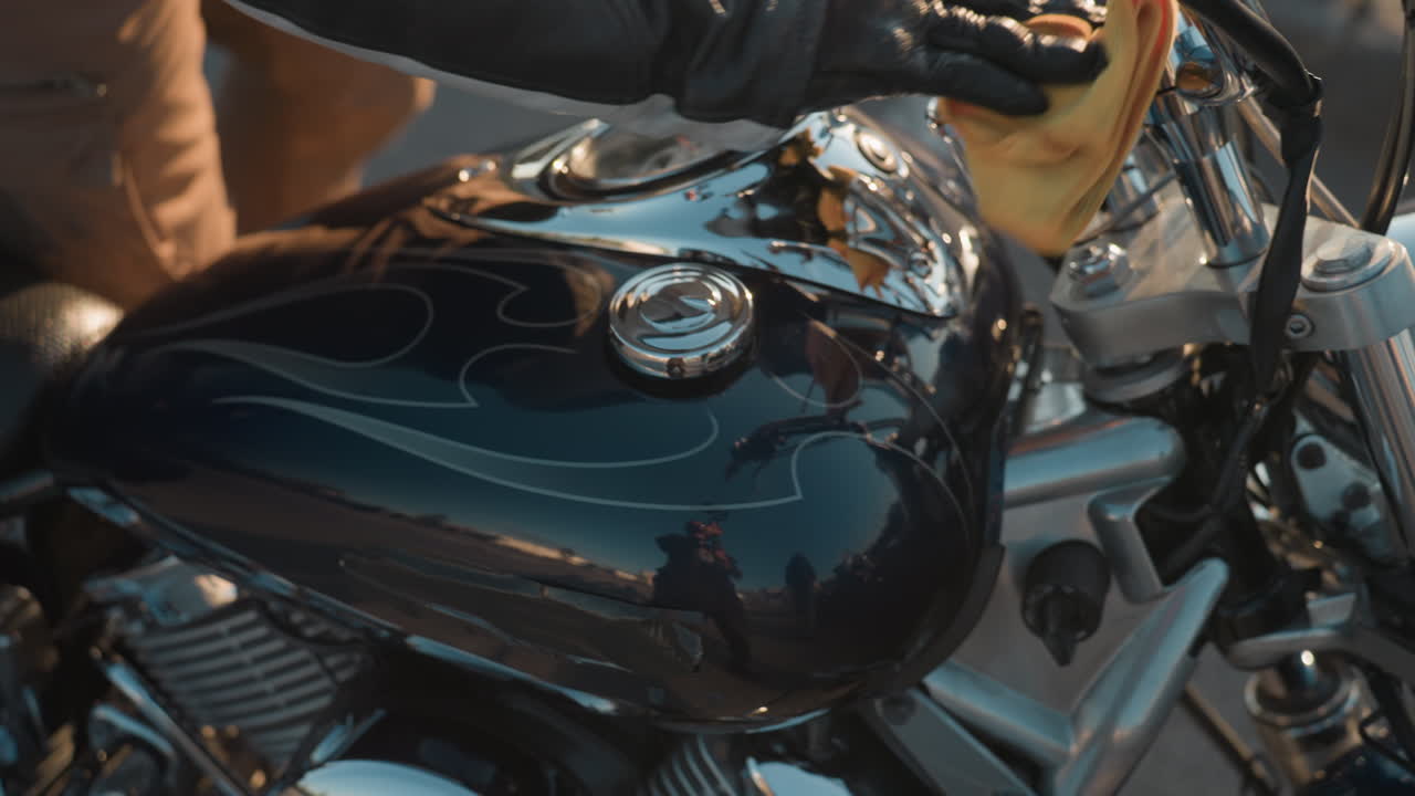 Rider wearing leather jacket adjusts gloves while holding yellow cloth outdoors, preparing for motorcycle ride under clear sky with blurred background of cars and urban setting in soft daylight