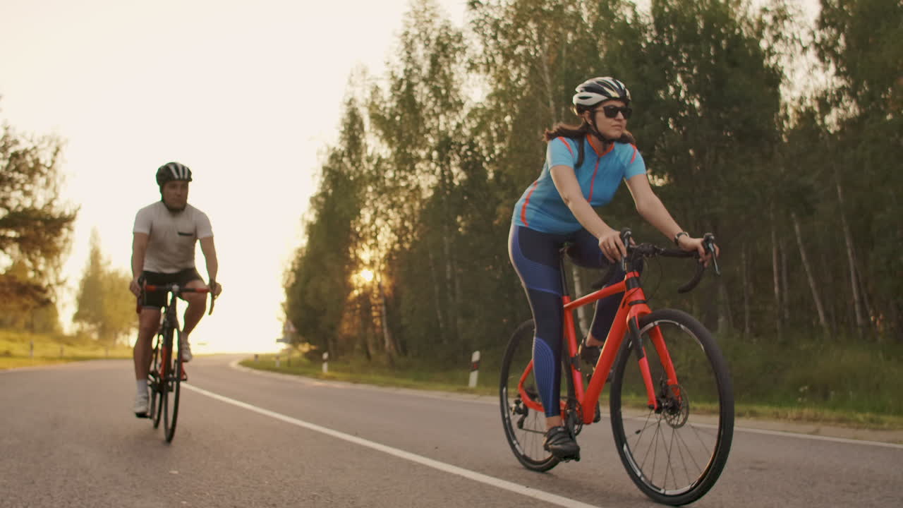 Two cyclists a man and a woman ride on the highway on road bikes wearing helmets and sportswear at sunset in slow motion
