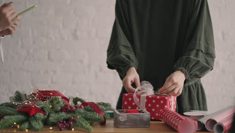 Crop-hands-decorating-wrapped-box-with-string.-Female-packing-cardboard-gift-box-on-wooden-table-with-various-decorative-items-prepared-for-Christmas-celebration.-Detail-female-hands-tying