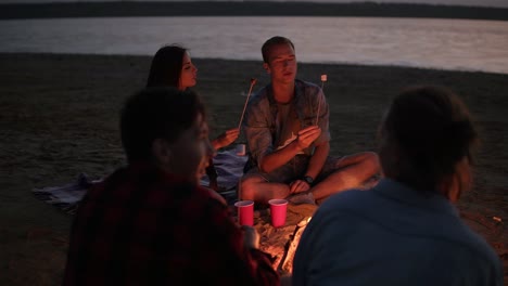 Young-couple-on-perspective-are-feeding-each-other-with-marshmallows-from-the-sticks.-Group-of-young-peopla-are-sitting-near-the-bonfire-on-the-beach.-Evening-dusk