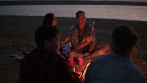 young couple on perspective are feeding each other with marshmallows from the sticks. group of young peopla are sitting near the bonfire on the beach. evening dusk