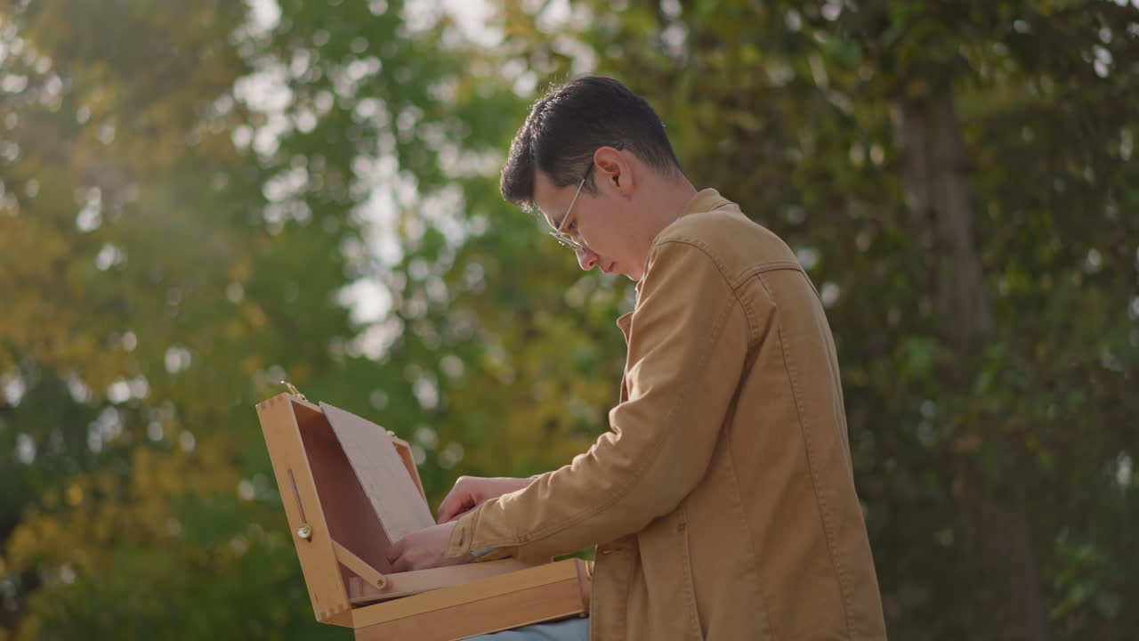 Pittore che dipinge durante l'ora d'oro in controluce, pittore assorto che lavora su aree di lavoro all'aperto con una calda illuminazione e una leggera luce soffusa sullo sfondo di una foresta, giacca casual, mani concentrate che aggiungono texture, atmosfera tranquilla e riflessiva