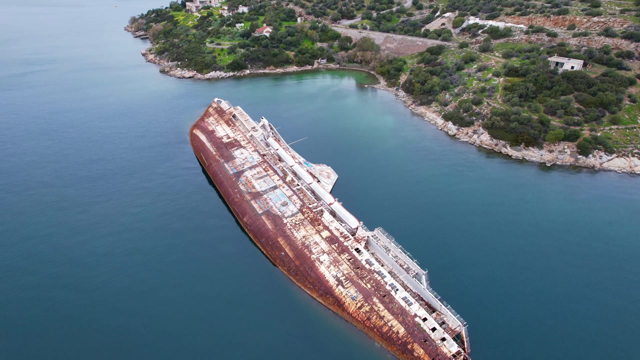 Aerial view of a large, rusty shipwreck lying on its side in a tranquil bay in greece