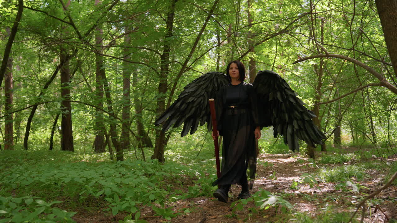Actress dressed as black fairy with large dark wings walks elegantly through green forest holding staff, sunlight glowing through trees, creating magical cinematic atmosphere
